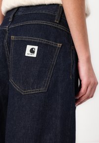 Tmavě modré džíny z denimu s žlutým prošíváním a bílou nášivkou na zadní kapse s logem. Džíny mají rovný střih a boční kapsy.