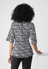 Schwarz-weißer, zebra gestreifter Oberteil mit Dreiviertelärmeln und abgerundetem Saum, der ein Schlüsselaugen-Detail am Rücken aufweist. Getragen mit schwarzen Hosen.