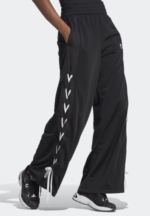 Pantalon de jogging noir à jambes larges avec un détail de lacets blancs sur les côtés, taille élastique, main dans la poche, porté avec des baskets noires et blanches.