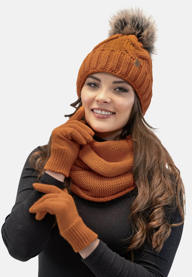 Strikket orange hue med en pom-pom, matchende handsker og et struktureret tørklæde, alt sammen i et varmt, hyggeligt stof. Handskerne har ribbede manchetter.