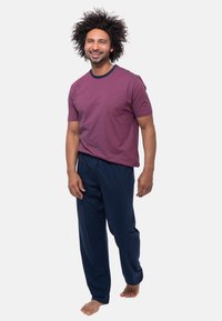 Glunderende man met krullend haar draagt een gestreept wijnrood T-shirt en marineblauwe broek, staat blootsvoets tegen een effen witte achtergrond.