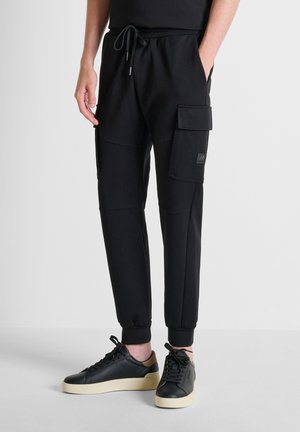 Uomo che indossa pantaloni jogger cargo neri con tasche laterali e sneakers nere con suola beige su sfondo chiaro.
