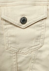 Beige bomullstyg med en spetsig ficka med klaff, som har en svart metallknapp och vertikala sömmar.