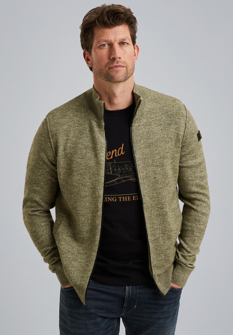 Grøn zip-up trøje med et tekstureret stof, stående krave og tætsiddende design over en sort t-shirt med grafisk tryk.