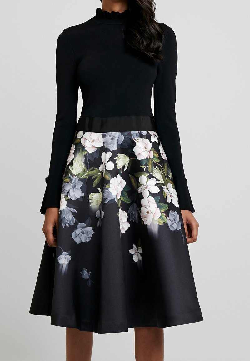 Zwarte longsleeve top met rimpelstaande kraag, gecombineerd met een klokrok met een bloemenprint in wit en lichtblauw op een donkere achtergrond.