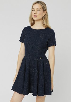 Robe bleu marine en tissu texturé avec des fils dorés, manches courtes, encolure ronde et jupe évasée avec poches avant.