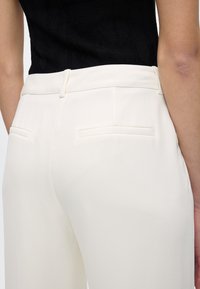 Κοντινό πλάνο ενός ατόμου που φοράει λευκό παντελόνι με ψηλή μέση και πίσω τσέπες και μια μαύρη υφή αμάνικη μπλούζα.