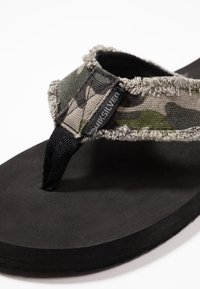 Infradito con motivo mimetico, caratterizzati da una suola in gomma nera e una fascia in tela sfilacciata con un'etichetta "Quiksilver". La fascia testurizzata aggiunge un ulteriore tocco di dettaglio.