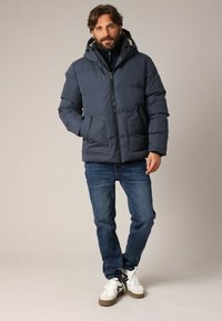 Doudoune bleu foncé avec capuche, présentant une fermeture éclair à l'avant et deux poches latérales, portée par-dessus un pull noir et un jean bleu.