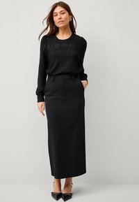 Haut en dentelle noire à manches longues avec un design texturé, associé à une jupe crayon noire taille haute. Des escarpins noirs à bouts pointus complètent la tenue.