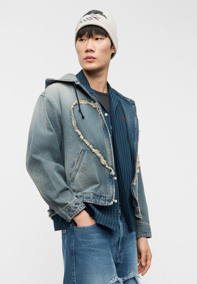 Νέος άνδρας που φοράει μπεζ σκουφάκι, μια φθαρμένη κοντή τζιν μπουφάν πάνω από ένα μπλε καρό πουκάμισο, και σκισμένο τζιν παντελόνι, στέκεται με το ένα χέρι στην τσέπη.