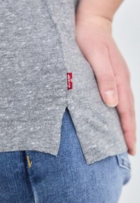 Pilkas medvilnės marškinėliai su šiek tiek tekstūros, turintys šoninį plyšį ir raudoną Levi's etiketę arti apdailos, derinami su mėlynomis džinsinėmis kelnėmis.