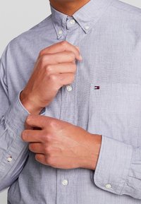 Vaaleanharmaa napillinen kauluspaita, jossa on tasku, valkoiset napit ja pieni kolmionmuotoinen logodetaili rinnassa. Kangas näyttää teksturoidulta.