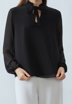 Vrouw draagt een zwarte blouse met lange mouwen, gerimpelde kraag en strik, gecombineerd met een witte broek, staand tegen een lichte achtergrond.