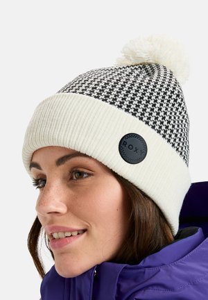 Frau trägt eine schwarz-weiße Hahnentritt-Strickmütze mit weißem Bommel und eine lila Winterjacke, lächelt sanft.