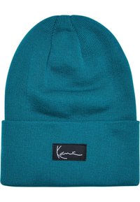 Teal színű kötött sapka hajtott peremmel. Fekete téglalap alakú címkével rendelkezik, fehér aláírás varrással. Puha tapintású, rugalmas anyag.