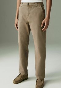 Pantalons en coton beiges avec une coupe droite, dotés d'une fermeture à bouton unique et de poches latérales, associés à des chaussures en daim marron.