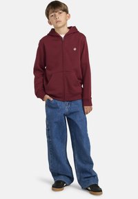 Sudadera con cremallera en color marrón burdeos, con bolsillos frontales y capucha, combinada con unos pantalones vaqueros azules de corte holgado. El conjunto presenta un diseño casual.