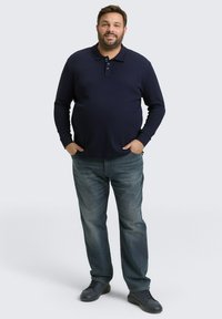 Marineblauwe polo met lange mouwen en drie-knopen sluiting, gecombineerd met vervaagde blauwe jeans en donkere sneakers, staand op een neutrale achtergrond.