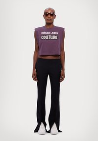 Lilla kort tank topp med teksten "VERSACE JEANS COUTURE" i hvit; kombinert med sorte tettsittende bukser med splitter, og hvite joggesko.