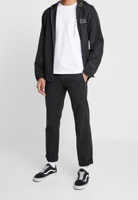 Svart lättviktig jacka med dragsko i huvan, vit t-shirt, svarta byxor och svarta sneakers med vita accenter. Enkelt design och avslappnad passform.