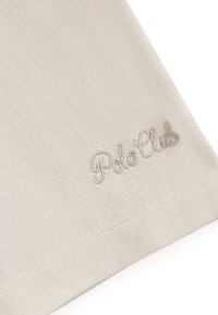 Beige stof met een gestructureerde weving, met zilver geborduurde "Polo Club" tekst aan één kant. Randen zijn gestikt voor duurzaamheid.