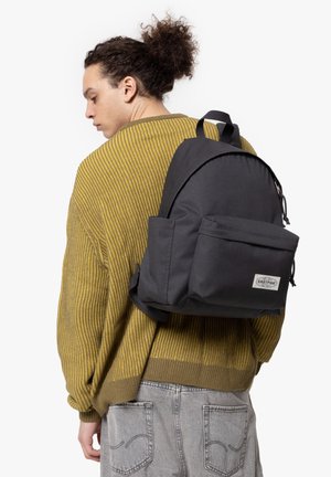 Zaino nero realizzato in tessuto resistente, con una forma arrotondata, tasca frontale e spallacci regolabili, indossato sopra un maglione giallo a trama.