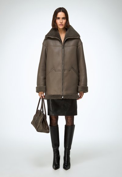 Chaqueta de piel marrón con forro de borrego, cuello alto, cierre frontal con cremallera y ribete de piel. Combinada con una falda de cuero negra y botas negras hasta la rodilla.