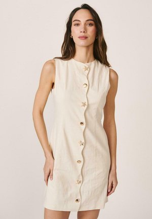 Mujer con cabello largo y castaño, vistiendo un vestido beige sin mangas con parte delantera ondulada y botones en forma de estrella, de pie frente a un fondo liso y claro.