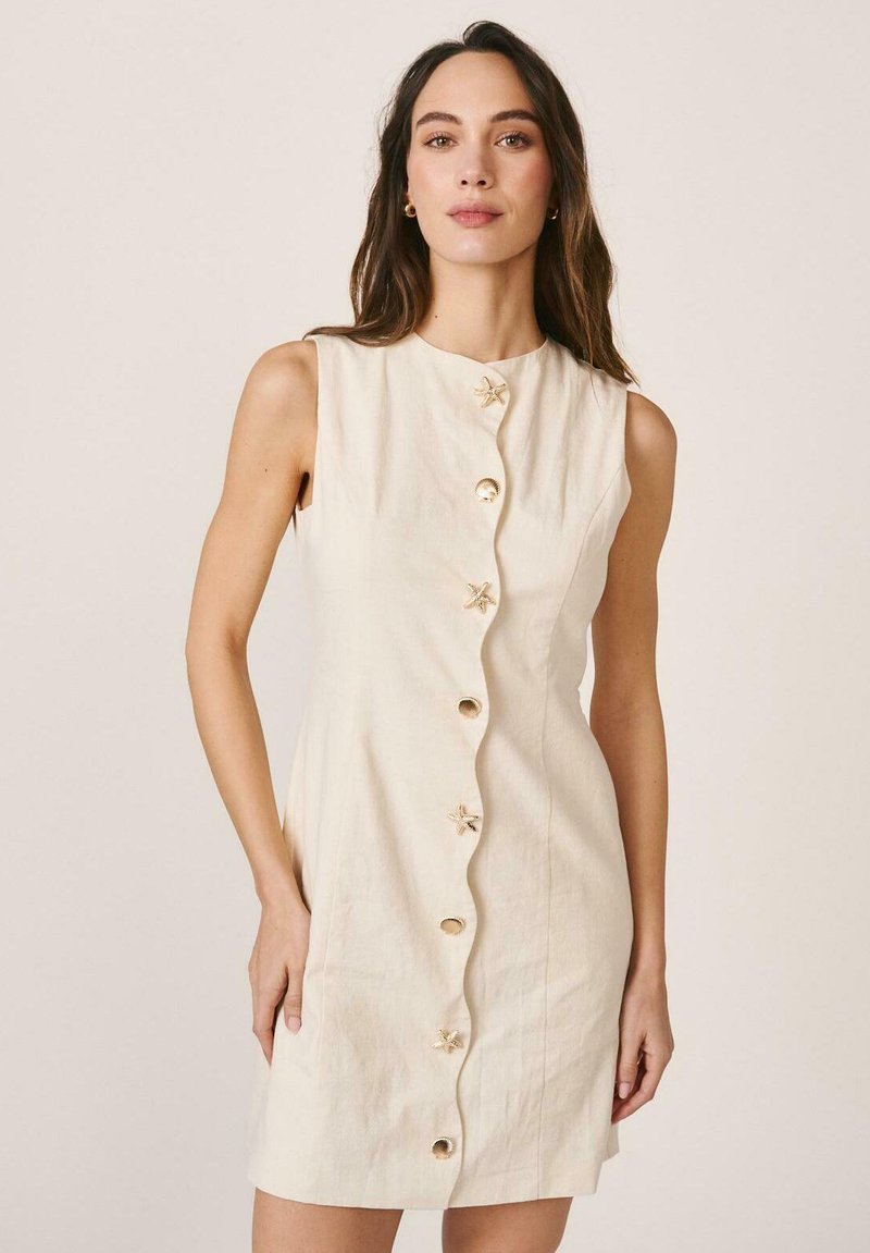 Mujer con cabello largo y castaño, vistiendo un vestido beige sin mangas con parte delantera ondulada y botones en forma de estrella, de pie frente a un fondo liso y claro.