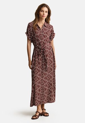Frau trägt ein braunes Maxikleid mit weißen abstrakten kreisförmigen Mustern, kurzen Ärmeln, Kragen, Taillengürtel und braunen Sandalen mit niedrigen Absätzen.