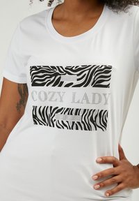 Camiseta de manga corta blanca con un patrón de rayas de cebra en blanco y negro, con "COZY LADY" en letras plateadas brillantes.