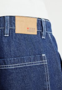 Jeans iz denima s svetlo rjavim usnjenim označevalcem, na katerem je napis "DENIM PROJECT". Imajo modro tkanino, bele šive in zasnovo zadnjega žepa.