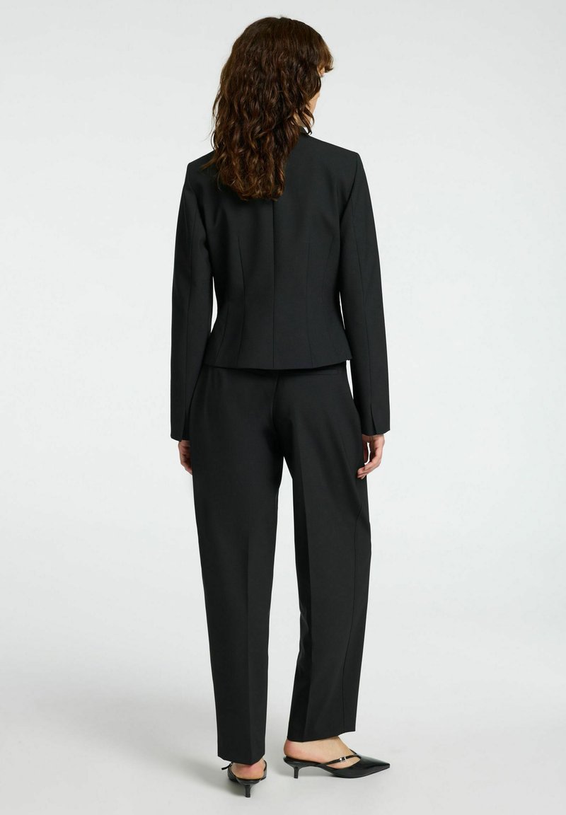 Schwarzer, taillierter Anzug mit einer eng anliegenden Jacke und weit geschnittenen Hosen. Besitzt eine glatte Textur und ein minimalistisches Design; enthält kleine schwarze Absätze.