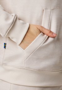 Tessuto di felpa beige chiaro con tasca a marsupio. Polsini e orlo a coste testurizzate, con un piccolo accento a bandiera blu e gialla.