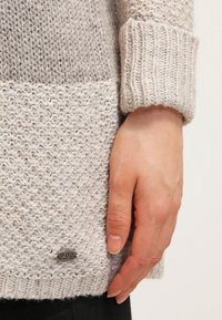 Pull en maille de couleur claire avec un motif texturé, des poignets côtelés et une petite étiquette métallique au niveau de l'ourlet. Apparence confortable et chaude.