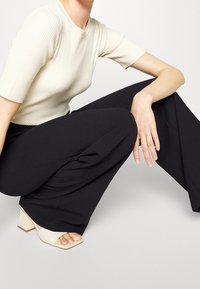 Kobieta w pozycji klęczącej, ubrana w białą koszulkę z krótkim rękawem o prążkowanej fakturze, czarne szerokie spodnie oraz beżowe sandały na obcasie z odkrytymi palcami, na neutralnym tle.