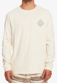 Beige långärmad skjorta i mjukt tyg, med en liten logotyp i diamantform på vänster bröst och ribbade ärmslut.