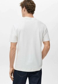 Moški z kodrastimi lasmi, oblečen v preprost beli kratkotrajni majici s kratkimi rokavi in temnimi kavbojkami, stoji obrnjeni stran od bele ozadja.