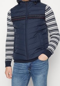 Marineblauer Puffer-Weste mit horizontalen Streifen und Kapuze, kombiniert mit einem langärmligen, gemusterten Strickshirt mit geometrischem Design.