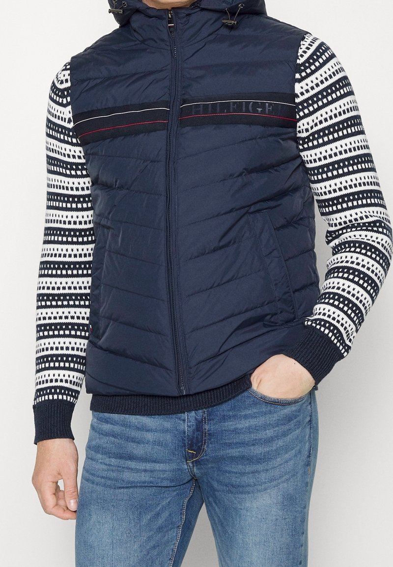 Marineblauer Puffer-Weste mit horizontalen Streifen und Kapuze, kombiniert mit einem langärmligen, gemusterten Strickshirt mit geometrischem Design.