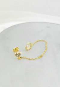 Orecchino color oro con un charm a forma di cuore ornato da tre strass trasparenti, collegato da una delicata catena a un piccolo orecchino con cuffia.