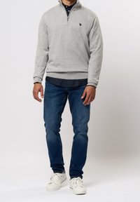 Man wearing light grey quarter-zip sweater, navy shirt, blue jeans, and white sneakers standing against a plain white background.