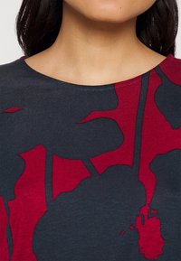 Haut marine avec un grand imprimé floral rouge. Présente un col rond et des manches courtes. Tissu doux et lisse avec une surface légèrement texturée.