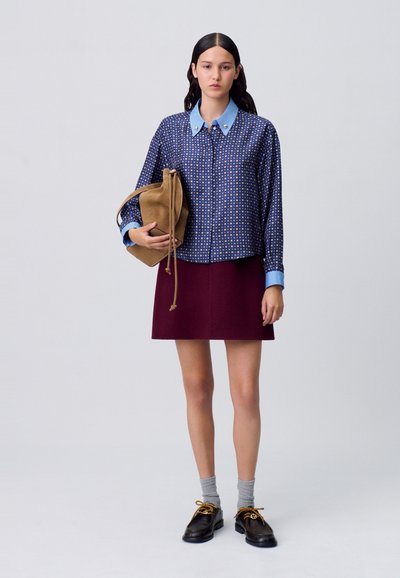 Camisa de botones con estampado azul marino y cuello azul claro, combinada con una falda burdeos. El modelo sostiene un bolso de ante beige, lleva calcetines grises y zapatos negros.