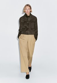 Hnědá semišová bunda se dvěma předními kapsami, nošená přes olivový rolák. Kombinována s krémovými širokými kalhotami a černými mulemi, stojící před bílým pozadím.