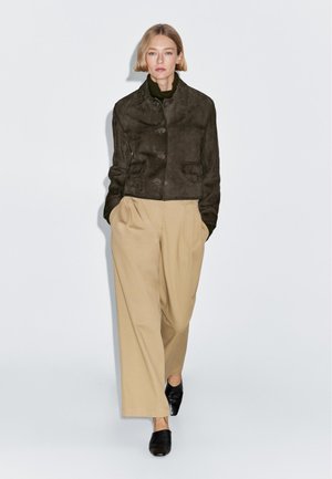 Hnědá semišová bunda se dvěma předními kapsami, nošená přes olivový rolák. Kombinována s krémovými širokými kalhotami a černými mulemi, stojící před bílým pozadím.