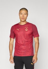 Piros, rövid ujjú mez, amely finom mintázattal rendelkezik, arany logóval és díszítésekkel. Könnyű anyag, kerek nyakú dizájn és testhezálló fazon.