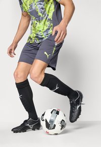 Μαύρα ποδοσφαιρικά παπούτσια, γκρι σορτσάκι με νεον πράσινες λεπτομέρειες, φανέλα με σχέδιο που συνδυάζει γκρι και πράσινο, μαύρες κάλτσες μέχρι το γόνατο, κοντά μια μπάλα ποδοσφαίρου.