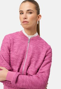 Veste bomber rose texturée avec une fermeture éclair à l'avant, poignets et col côtelés. Le tissu présente un motif bosselé pour plus de dimension.
